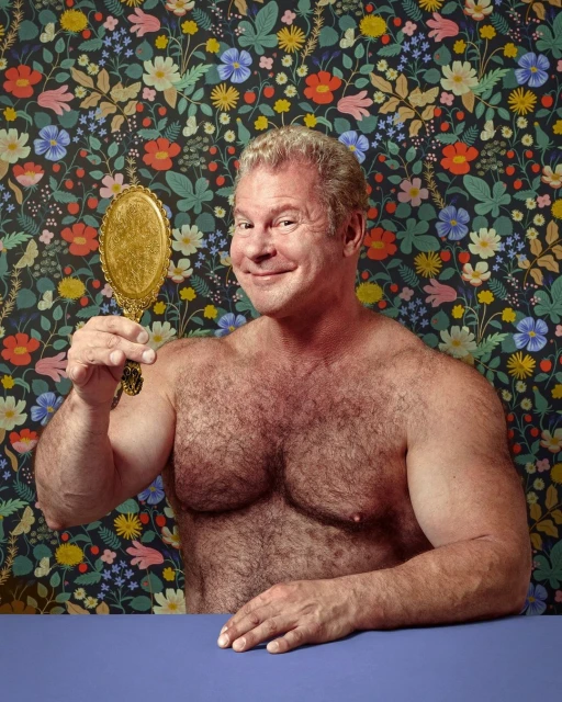 Adult man smiles while looking in a vintage gold hand mirror with a floral patterned background and blue table.